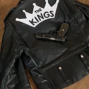 May include: Black leather jacket with the text "THE KINGS" in white, above a handgun. The jacket has a zipper closure and silver buttons. The jacket is hanging on a hook.