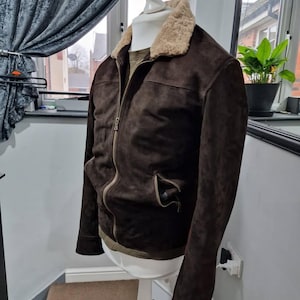 Op de afbeelding: Een donkerbruine su&egrave;de jas met een beige schapenvachtkraag en een ritssluiting. De jas heeft twee voorzakken en wordt gepresenteerd op een witte paspop. Medium maat.