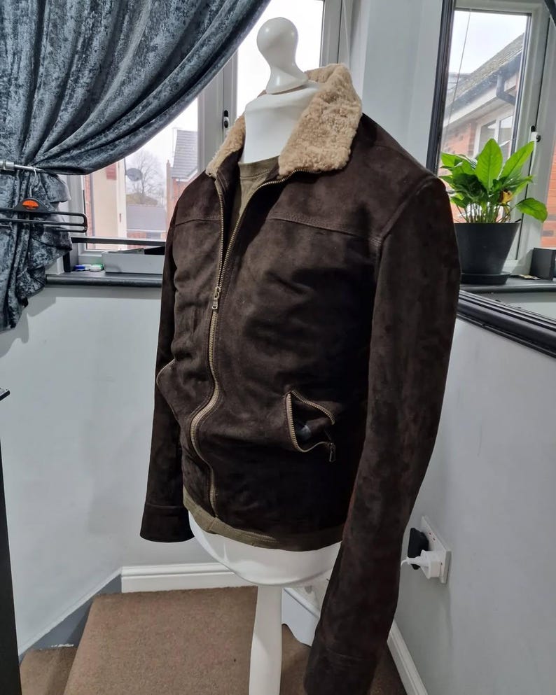 Op de afbeelding: Een donkerbruine su&egrave;de jas met een beige schapenvachtkraag en een ritssluiting. De jas heeft twee voorzakken en wordt gepresenteerd op een witte paspop. Medium maat.