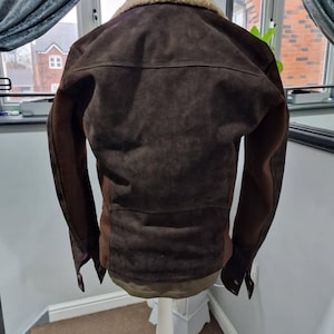 Op de afbeelding: Bruine su&egrave;de jas met een beige schapenvachtkraag. De jas heeft een klassiek ontwerp met een knoopsluiting en een getailleerde pasvorm. De mouwen zijn donkerbruin. De jas wordt op een witte paspop gepresenteerd.