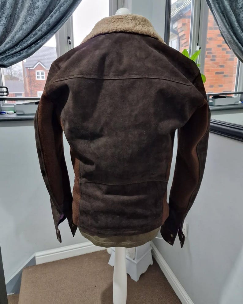 Op de afbeelding: Bruine su&egrave;de jas met een beige schapenvachtkraag. De jas heeft een klassiek ontwerp met een knoopsluiting en een getailleerde pasvorm. De mouwen zijn donkerbruin. De jas wordt op een witte paspop gepresenteerd.