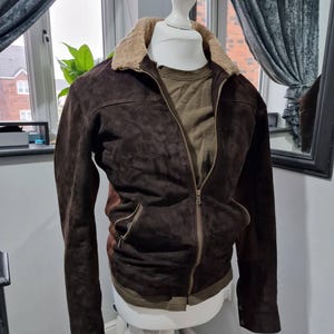 Op de afbeelding: Een bruine su&egrave;de jas met een beige imitatiebontkraag en een ritssluiting. De jas heeft twee voorzakken en wordt gepresenteerd op een witte paspop. Een groen shirt is eronder zichtbaar.