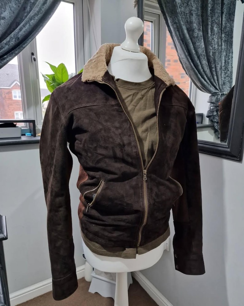 Op de afbeelding: Een bruine su&egrave;de jas met een beige imitatiebontkraag en een ritssluiting. De jas heeft twee voorzakken en wordt gepresenteerd op een witte paspop. Een groen shirt is eronder zichtbaar.