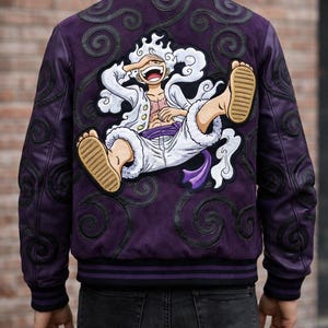 May include: Purple bomber jacket with a large cartoon graphic on the back. The jacket features a character in white clothing with a purple sash and brown sandals. Black swirl designs and trim accent the jacket.