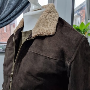 Op de afbeelding: Een donkerbruine su&egrave;de jas met een beige schapenvachtkraag. De jas heeft een ritssluiting en een klassiek ontwerp. De jas wordt op een mannequin getoond. Een stijlvolle buitenkleding.