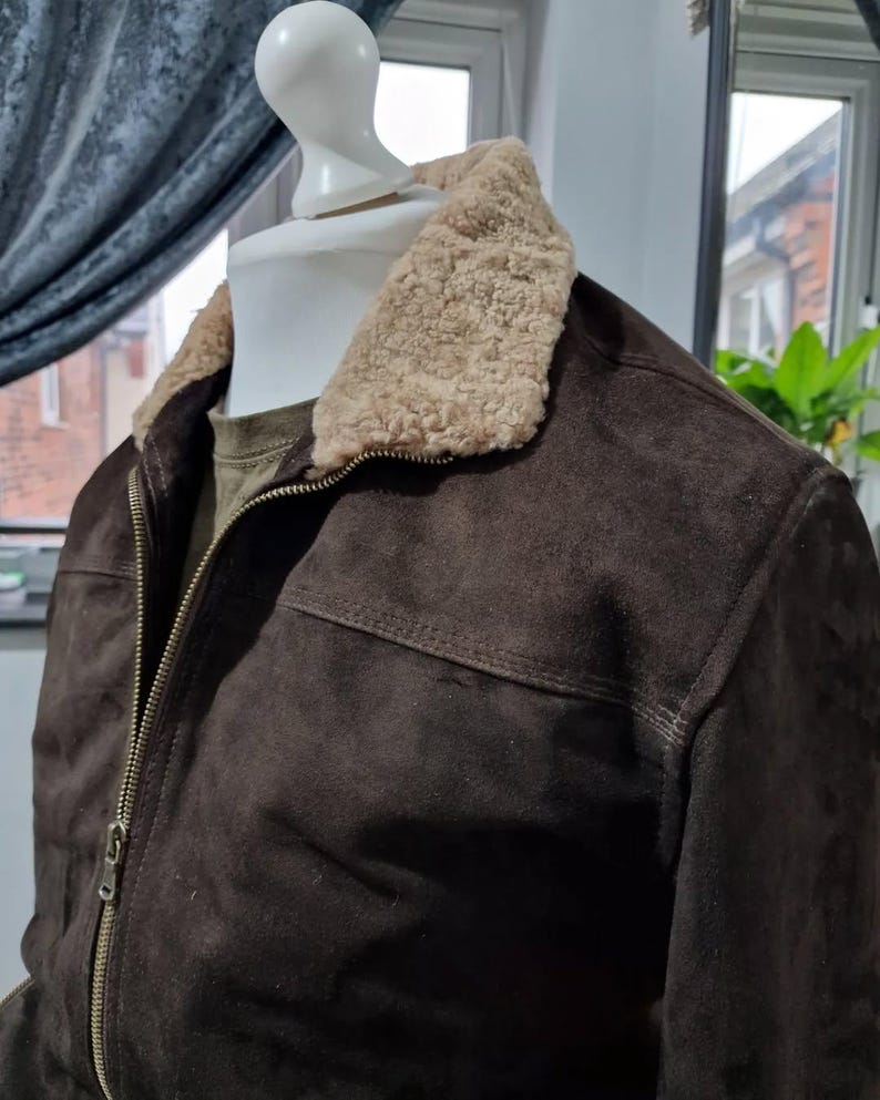 Op de afbeelding: Een donkerbruine su&egrave;de jas met een beige schapenvachtkraag. De jas heeft een ritssluiting en een klassiek ontwerp. De jas wordt op een mannequin getoond. Een stijlvolle buitenkleding.