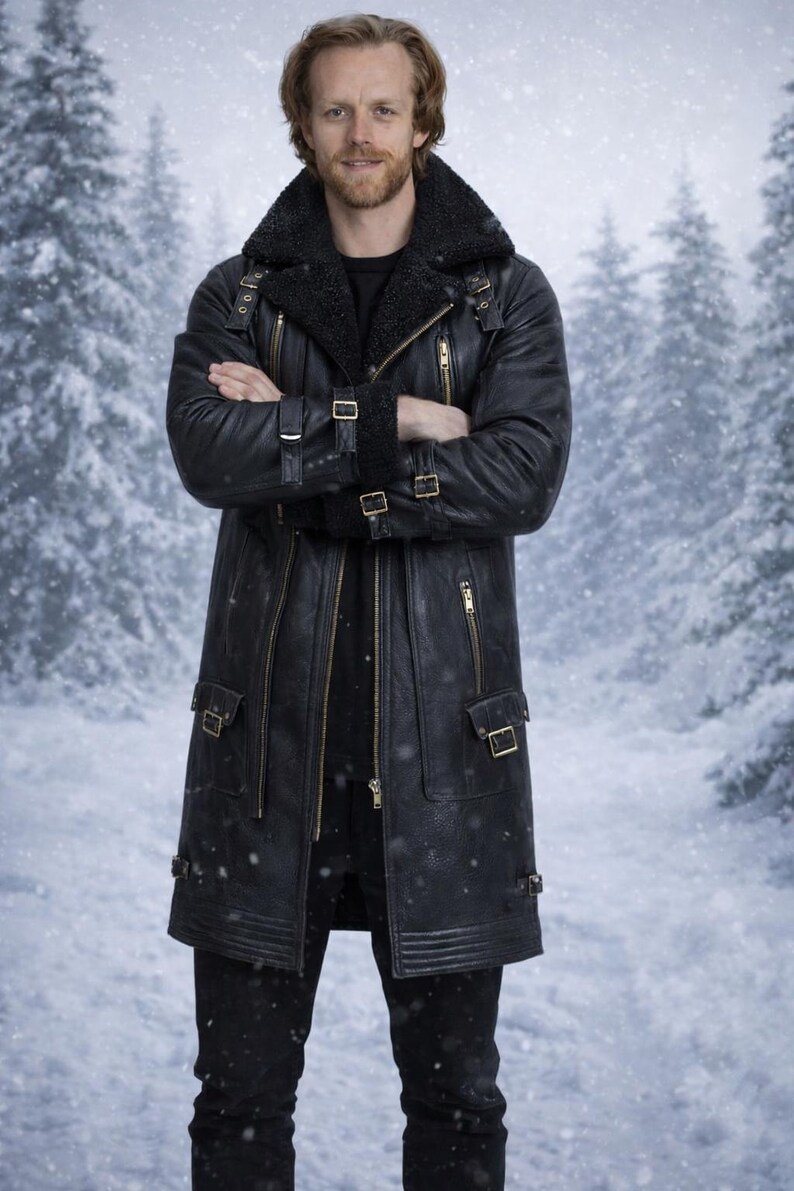 May include: A black leather coat with a shearling collar, multiple zippers, and buckle details. The long coat features a tailored fit and is worn over a black outfit. The background shows a snowy forest scene.