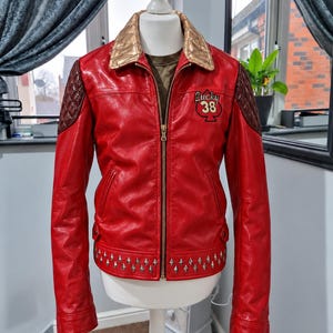 May include: A red leather jacket featuring a gold quilted collar, dark brown quilted shoulder panels, and a gold front zipper. The chest displays a black spade emblem with the text Lucky 38. The hem is decorated with a row of silver metal studs.