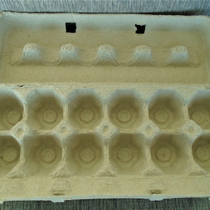 May include: An open, empty egg carton made of light brown recycled paper. The carton has two rows of six circular indentations designed to hold eggs. Two square cutouts are visible on the top half of the carton.