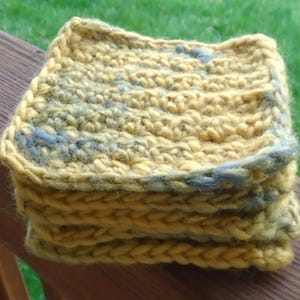 May include: A stack of four square, hand-crocheted coasters. The coasters are a mix of yellow and blue-grey yarn, with a thick, textured stitch. The coasters are sitting on a wooden surface, with green grass in the background.