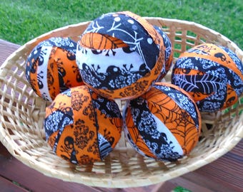May include: A woven basket filled with six fabric balls. The balls are decorated with Halloween-themed patterns in orange, black, and white. Designs include skulls, spiderwebs, and damask patterns. The basket sits on a wooden surface with green grass in the background.