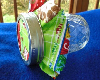 May include: A decorative jar lid with a fabric wrap. The lid is silver with a green and red patterned fabric. The jar is clear glass with a diamond pattern. The fabric wrap has a green and white plaid pattern.