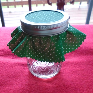 May include: A clear glass jar with a silver lid and a green fabric cover with white polka dots. The jar is sitting on a red surface.