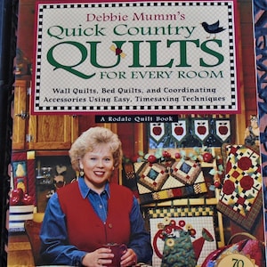 May include: Book cover featuring a woman holding a mug in a country kitchen setting. The book title is "Debbie Mumm's Quick Country Quilts for Every Room". The book cover also features a bird and a quilt with an apple design.