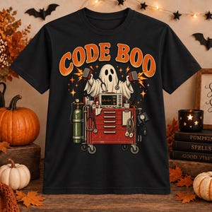 May include: A black t-shirt featuring a graphic of a ghost dressed as a medical professional standing behind a red medical cart. The ghost holds two defibrillator paddles. The text CODE BOO is printed in orange above the ghost. The design includes medical equipment like a monitor, oxygen tank, and stethoscope.