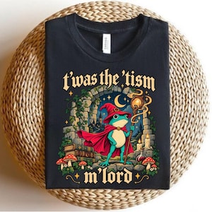 Può includere: Una t-shirt nera con la grafica di una rana verde che indossa un cappello e un mantello da mago rossi, mentre tiene in mano un bastone luminoso. Il design include archi in pietra, libri, funghi e candele, con il testo 't'was the 'tism m'lord'.