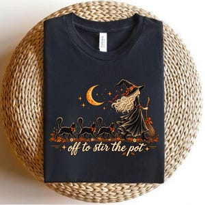 May include: A black t-shirt featuring a graphic of a witch with long hair wearing a hat and holding a broom, followed by three black cats. Above them is a crescent moon and stars. The text below reads, off to stir the pot.