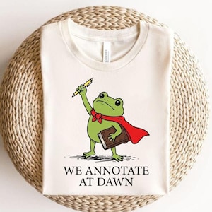May include: A cream-colored t-shirt featuring a graphic of a green frog wearing a red cape, holding a yellow pencil and a brown book. Below the illustration, the text reads "WE ANNOTATE AT DAWN" in black, capitalized, serif lettering.