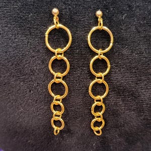May include: A pair of gold-colored earrings. Each earring features a small ball at the top, connected to a series of graduated gold rings. The rings are linked together, creating a dangling effect. The earrings are displayed against a dark background.