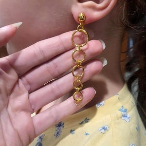 May include: Gold-colored dangle earrings featuring a series of connected circles. The earrings are shown worn on an ear, with a hand holding the lower part of the earrings. The person is wearing a yellow shirt with a floral pattern.