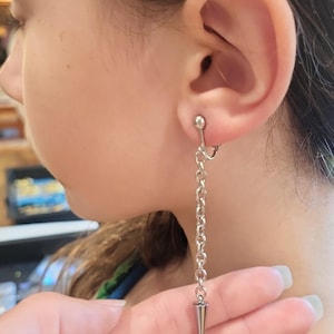 May include: Silver-toned clip-on earring featuring a chain and a conical charm. The earring is attached to the ear with a small ball-shaped clip. The chain hangs down, ending in a pointed cone. The earring is shown on a person's ear.