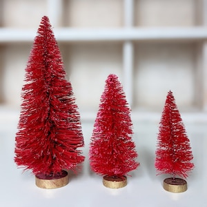May include: Three red bottle brush Christmas trees of varying sizes. The trees are on a white surface.