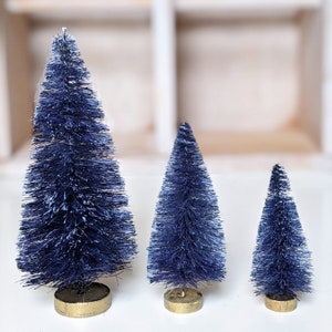 May include: Three blue bottle brush Christmas trees of varying sizes. The trees are on a white surface.