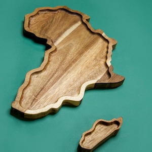 May include: Wooden serving tray and small dish, both shaped like the African continent. The tray has a natural wood grain finish with a carved-out interior. The small dish is a miniature version of the tray. The background is a solid teal colour.