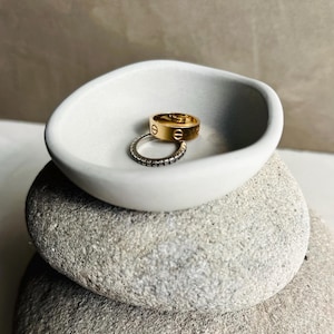 May include: A white, oval dish contains two rings: a gold band with engraved markings and a silver ring with small, dark stones. The dish sits on a stack of grey, smooth stones, creating a minimalist aesthetic.