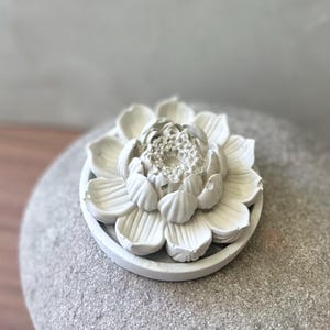 Op de afbeelding: Een wit, bloemvormig decoratief object. Het object heeft meerdere lagen bloemblaadjes en staat op een ronde, ondiepe basis. De bloemblaadjes zijn gedetailleerd met ribbels en het ontwerp is minimalistisch en elegant.