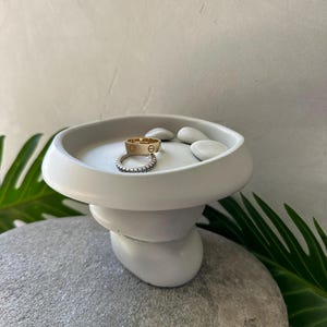 Peut inclure: Un porte-anneaux blanc à plusieurs niveaux avec un dessus rond. Deux bagues, une dorée et une argentée, reposent sur le dessus, ainsi que de petites pierres blanches lisses. Le support repose sur une grosse pierre grise, avec des feuilles vertes en arrière-plan.