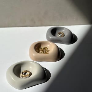 May include: Three organic-shaped ring dishes in neutral tones: light gray, beige, and dark gray. Each dish holds jewelry, including gold rings. The dishes are arranged in a row, with a shadow cast across the white surface.
