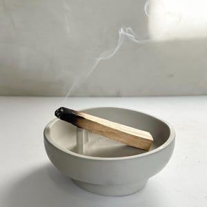 May include: A gray, round incense holder with a burning palo santo stick. The stick is light brown with a black, charred end, emitting wisps of white smoke. The holder is made of a smooth, matte material.