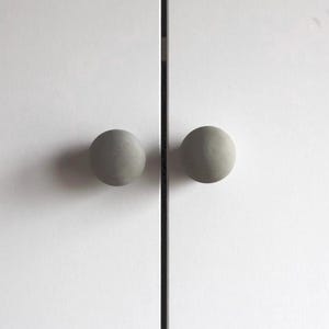 May include: Two gray, spherical cabinet knobs are mounted on a white cabinet door. The knobs are evenly spaced and have a smooth, matte finish. The cabinet doors are divided by a thin, black vertical line.