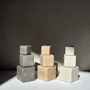 Può includere: Tre serie di blocchi quadrati impilati in varie tonalità neutre: grigio, beige e bianco sporco. Ogni serie è composta da tre blocchi di dimensioni decrescenti, creando una forma a piramide. I blocchi sono disposti su una superficie bianca.