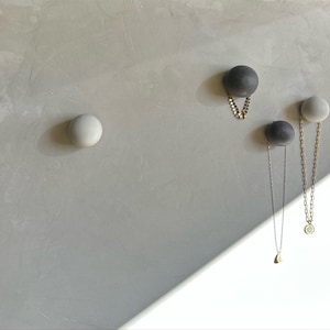 May include: A collection of decorative wall hooks in a minimalist style. The hooks are spherical, with two in a dark gray color and two in a light gray color. Two hooks are holding necklaces with gold and silver chains.