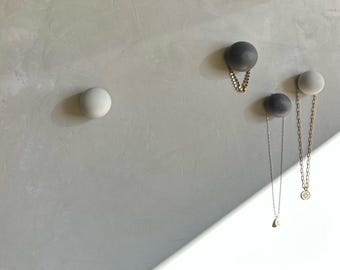 May include: A collection of modern wall hooks in shades of gray and white. Two dark gray hooks and one white hook are shown, each with a small chain or necklace hanging from them. The hooks are mounted on a light gray wall.