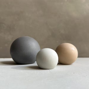 Puede incluir: Tres objetos decorativos esféricos de diferentes tamaños y colores. Una esfera grande de color gris oscuro a la izquierda, una esfera blanca más pequeña en el centro y una esfera de color beige claro a la derecha. Los objetos están sobre una superficie gris claro.
