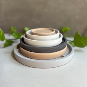 May include: A stack of five round, minimalist concrete trays in varying neutral colors: light gray, tan, white, dark gray, and a smaller tan tray on top. The trays are stacked, with the largest at the bottom. Green leaves are in the background.
