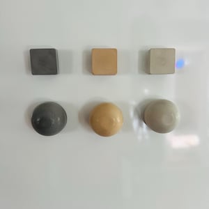 以下が含まれることがあります： さまざまな形と色をした6つの装飾用マグネットのコレクション。グレー、タン、オフホワイトの3つの正方形のマグネットが、グレー、タン、オフホワイトの3つの丸いマグネットの上に配置されています。マグネットは白い表面に配置されています。