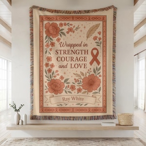 May include: A beige woven throw blanket with a floral design and the words "Wrapped in Strength Courage and Love." The blanket has a ribbon border and a fringed edge. The name "Ray White" is on a banner.