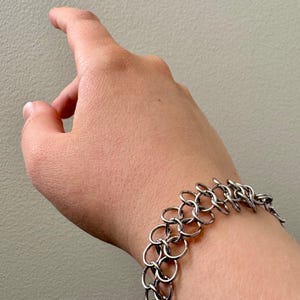 Könnte beinhalten: Ein silberfarbenes Kettenhemd-Armband aus mehreren ineinandergreifenden, kreisförmigen Metallringen. Das Armband weist ein zweireihiges Muster aus verbundenen Schlaufen auf, die sich um das Handgelenk legen und ein flexibles, strukturiertes Metallband bilden.