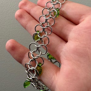 Könnte beinhalten: Ein Kettenarmband aus ineinandergreifenden silberfarbenen Metallringen. Das Armband ist mit kleinen, durchscheinenden grünen, blattförmigen Perlen verziert, die zwischen den Metallgliedern eingearbeitet sind und ein sich wiederholendes Muster bilden.