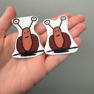 May include: Two whimsical snail-shaped patches. Each patch features a white background with a brown snail body and a light brown face. The snails have black antennae and a black line at the base. The patches are held in a hand.