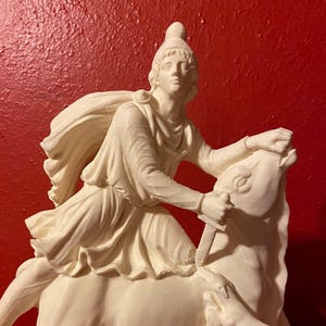 May include: A cream-coloured sculpture of a figure on horseback, wielding a sword. The rider wears a helmet and flowing robes. The horse rears, with a smaller animal beneath. The sculpture is set against a red background.
