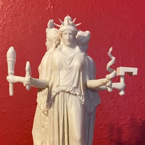 Può includere: Statua bianca di tre figure vestite, ognuna con oggetti simbolici. La figura centrale, incoronata da un diadema appuntito, è in evidenza. La statua è su una base rotonda, su sfondo rosso.