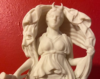 May include: A white statue of a figure wearing a draped garment, featuring small horns on the head and holding a cylindrical object in the right hand. The figure is positioned with fabric billowing behind the head and shoulders in a flowing manner.