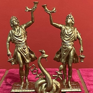 Könnte beinhalten: Ein Set aus drei goldfarbenen, metallischen Dekorationsobjekten. Zwei identische Figuren stehen aufrecht, jede hält ein hornförmiges Gefäß in die Höhe und einen kleinen Korb. Eine zentrale, gewundene Schlangenfigur zeigt zwei Symbole.