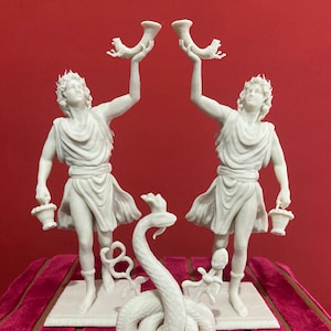 Könnte beinhalten: Zwei identische weiße Statuen von Figuren in drapierten Gewändern, die jeweils ein hornförmiges Gefäß in die Höhe halten und einen kleinen Korb an der Seite tragen. Dazwischen steht eine weiße, gewundene Schlangenskulptur mit einem Kamm.
