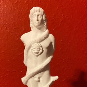 May include: A white statue of a male figure with a snake wrapped around the body. The figure has a lion's head on the chest. The statue is standing on a red surface.